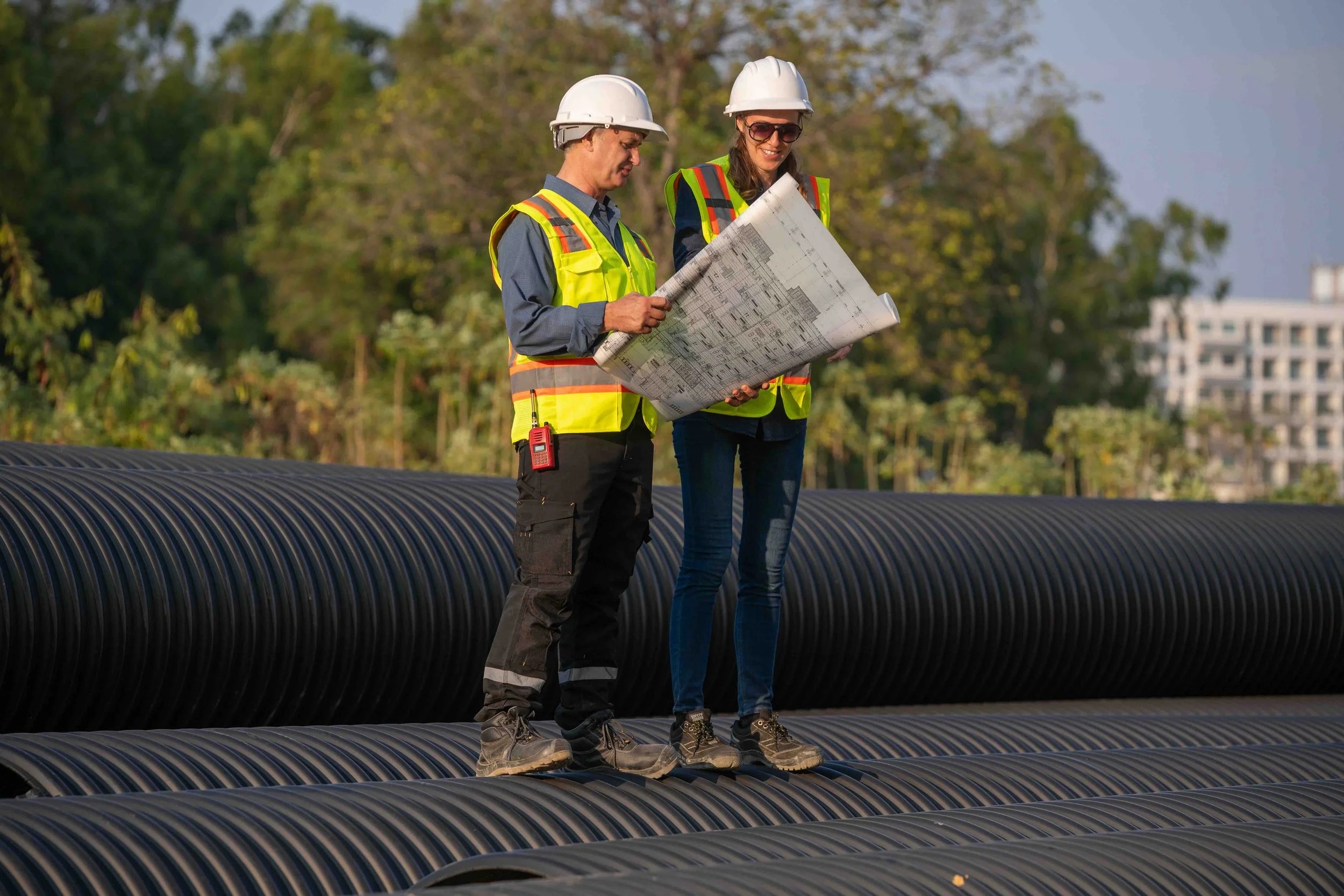 Two civil engineering professionals in hard hats and high-visibility vests standing on large corrugated pipes, actively reviewing construction blueprints on-site.